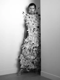 Model wearing a voluminous textured white petal gown and statement earrings in a white corner for a black-and-white fashion editorial.