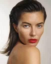 Close-up portrait of a woman with slicked-back wet hair and bold red lipstick looking over her bare shoulder against a pale backdrop.