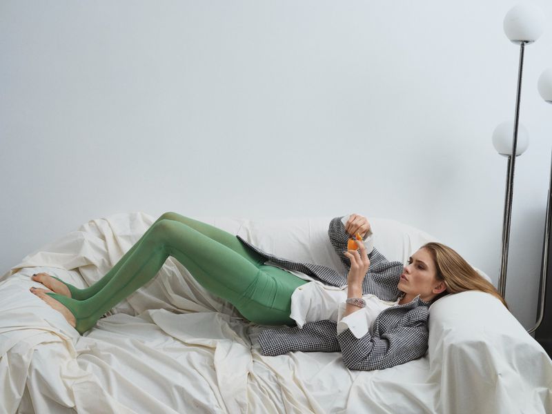 Woman lounges on white couch in green tights and patterned blazer, holding an orange; floor lamp stands in background.