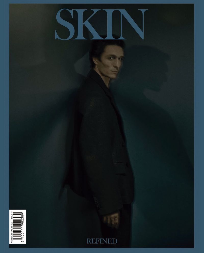 A man poses in a jacket for a magazine cover featuring the word 'SKIN' on a teal background, creating a fashionable editorial shot.