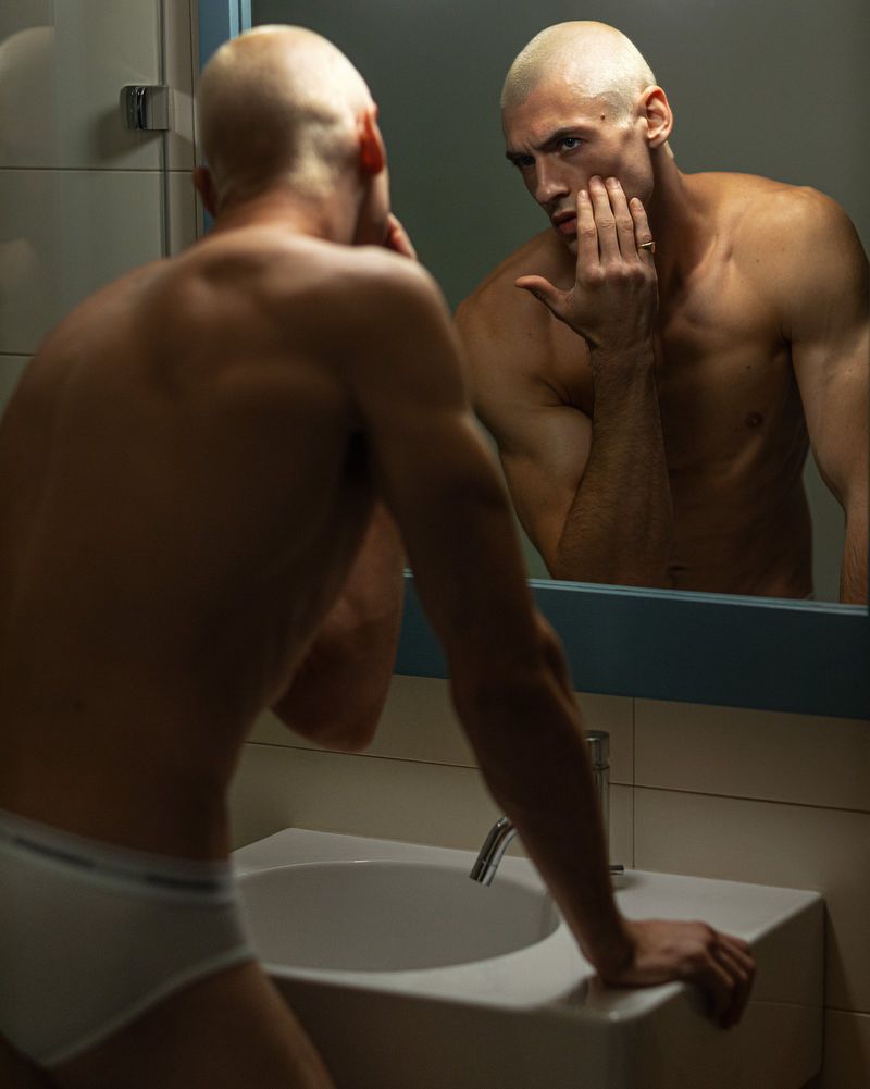 Muscular man with a shaved head stands shirtless at a bathroom sink, looking at his reflection in the mirror with his hand on his face.