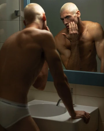 Muscular man with a shaved head stands shirtless at a bathroom sink, looking at his reflection in the mirror with his hand on his face.