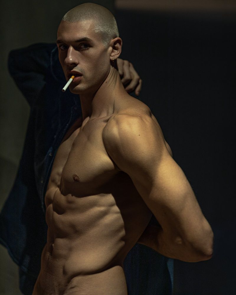 Shirtless male model with a cigarette poses indoors with dark background, showcasing his physique with dramatic lighting.