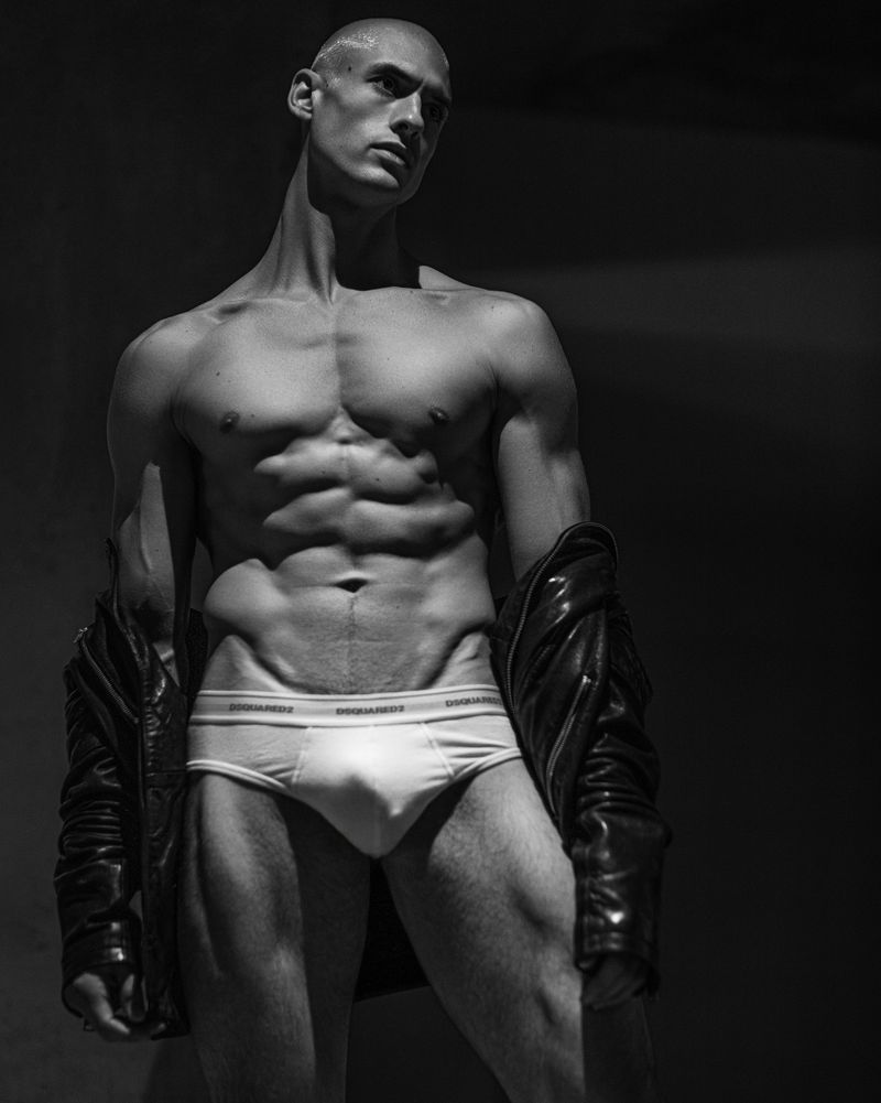 Black and white portrait of muscular adult male in underwear and leather jacket, posing in a photo studio with a dark background.