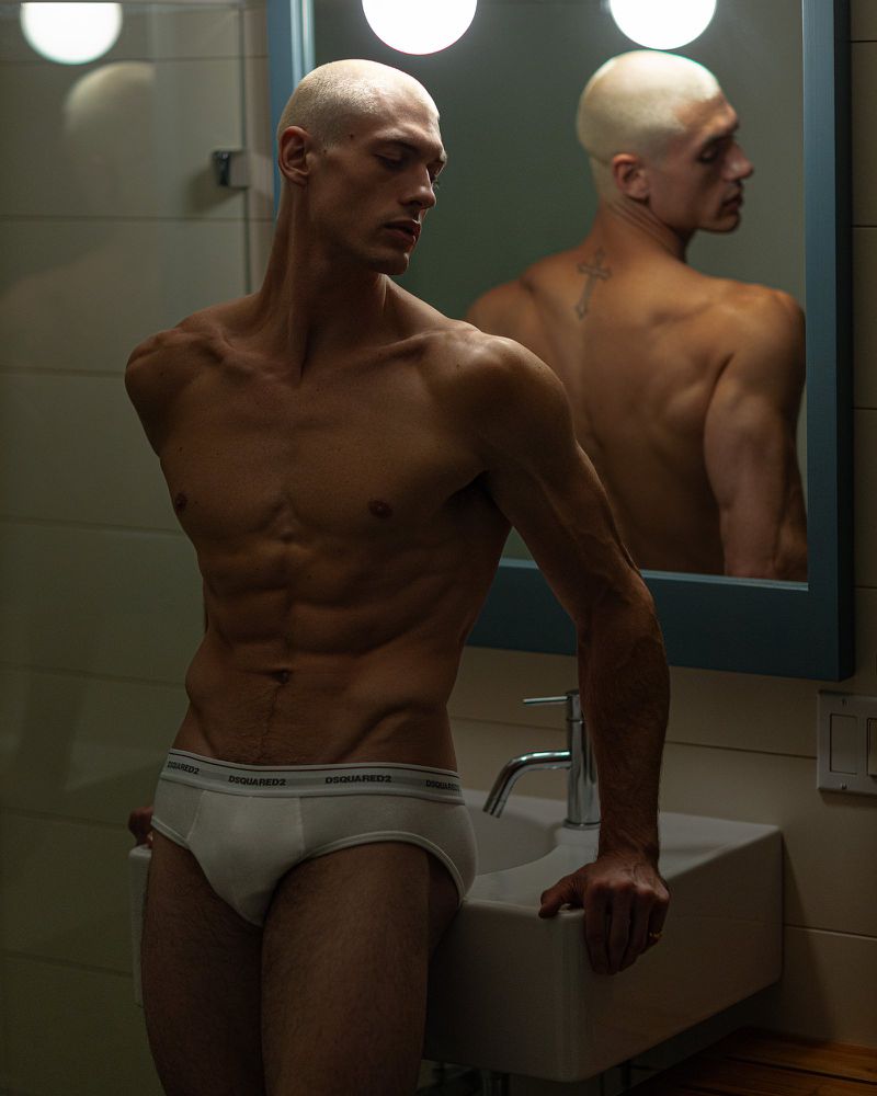 Shirtless man with shaved head stands in bathroom with white briefs, leaning on the sink, a mirror showing a reflection of his back.