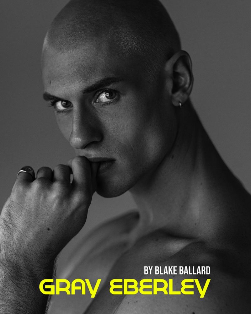 Portrait of a fair-skinned adult male with a shaved head, jewelry, neutral facial expression, and hand-to-mouth gesture, against a gray background.