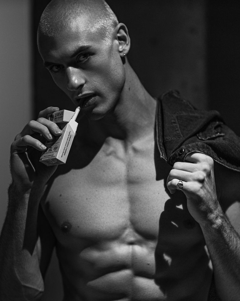 Black and white portrait of a muscular, bald man with a cigarette in his mouth, wearing a denim jacket, and posing indoors.