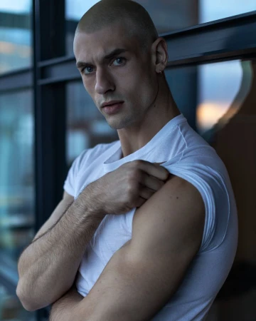 Close-up portrait of a bald male model wearing a white t-shirt, posing indoors near a dark window frame.