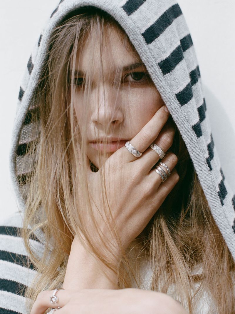 Close-up of a young adult with blonde hair wearing a striped hooded sweater and several rings, posing against a white backdrop.