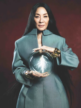 Michelle Yeoh holds a glowing orb wearing an oversized grey coat with black hair, gold jewelry, and long nails in a studio shot.