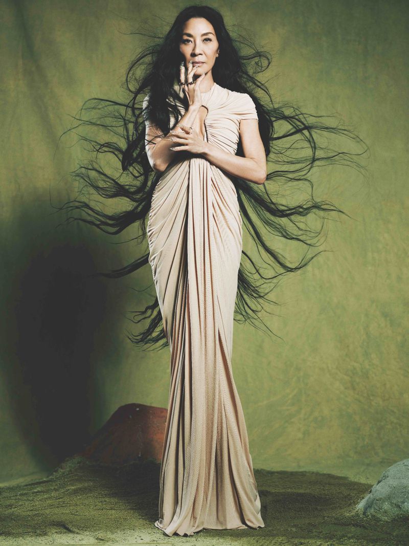 Michelle Yeoh poses in a floor-length, draped cream dress with long dark hair against a green background, her hands gracefully near her face.