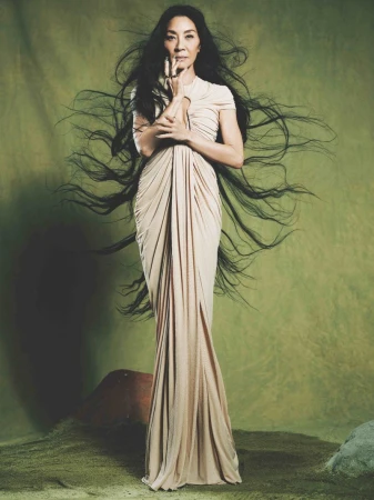 Michelle Yeoh poses in a floor-length, draped cream dress with long dark hair against a green background, her hands gracefully near her face.