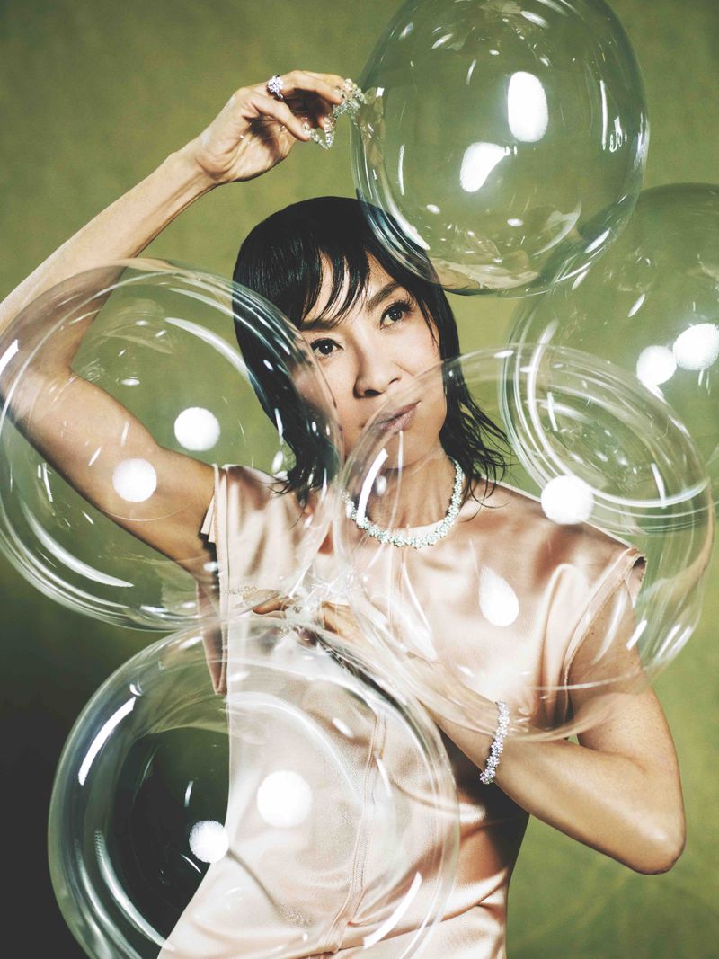 Michelle Yeoh portrait surrounded by transparent balloons, wearing a light pink dress and diamond jewelry against a green background.