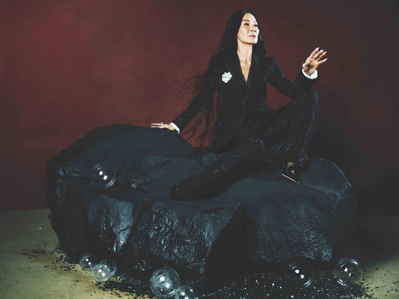 Michelle Yeoh poses in a black sequin suit on a black rock-like formation, surrounded by sparkling specks and glass balls against a rusty backdrop.