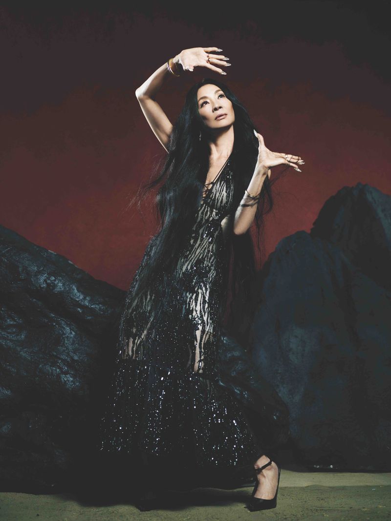 Michelle Yeoh strikes a pose in a dark semi-sheer gown against a dark red background with abstract rock-like structures in the studio.