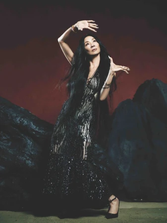 Michelle Yeoh strikes a pose in a dark semi-sheer gown against a dark red background with abstract rock-like structures in the studio.