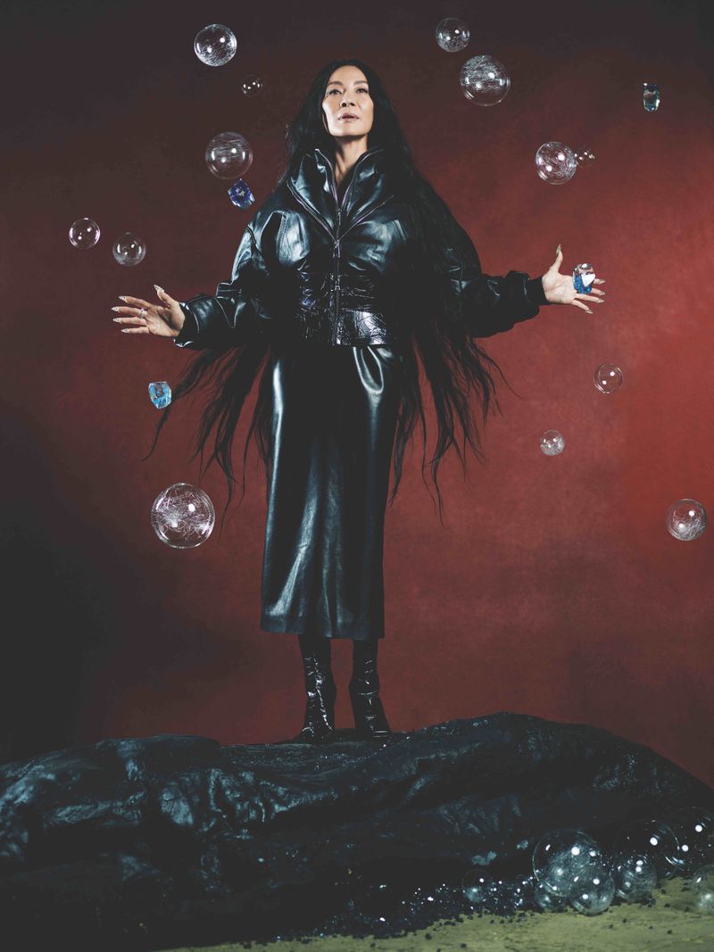 Elegant Asian woman in black leather outfit stands on a black rock surrounded by soap bubbles against a red-brown backdrop.
