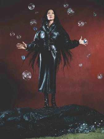 Elegant Asian woman in black leather outfit stands on a black rock surrounded by soap bubbles against a red-brown backdrop.