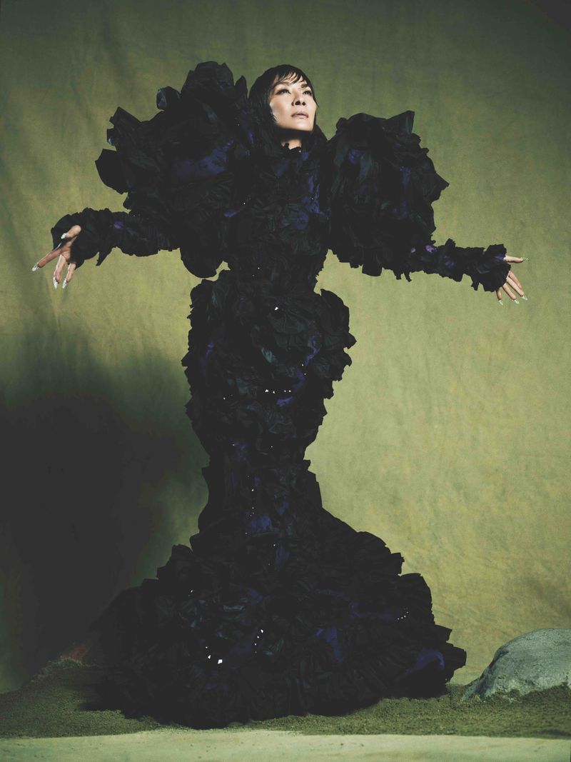 Dramatic full-length portrait of an adult woman in a sculptural ruffled black dress, against a soft green backdrop with natural stone.