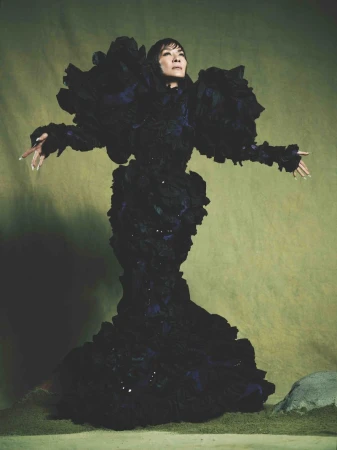 Dramatic full-length portrait of an adult woman in a sculptural ruffled black dress, against a soft green backdrop with natural stone.