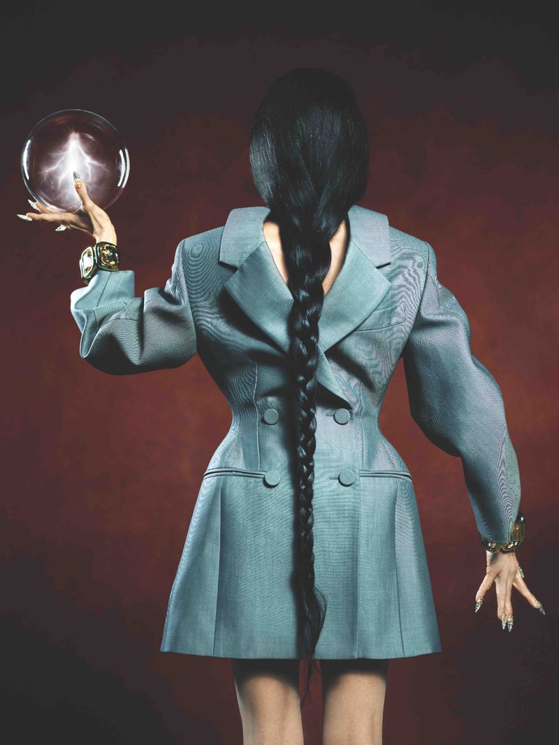 Woman with long braid poses in a structured blazer dress, holding a glowing orb, fashion-forward and artistic against a rich backdrop.