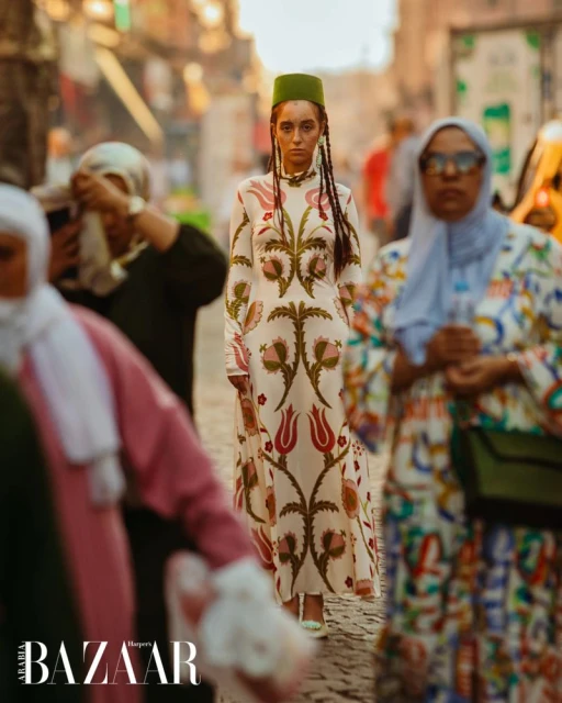 Fashion model wearing floral dress and green fez walks down city street with onlookers, photographed for Harper's Bazaar magazine.