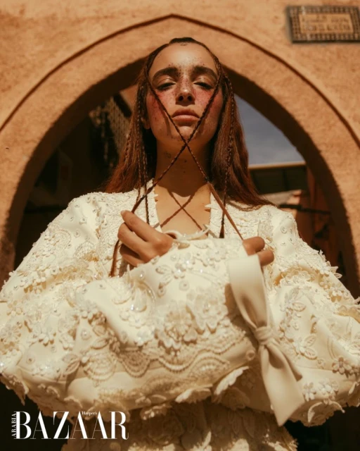 Portrait of a female model wearing a white lace dress, holding a floral lace bag, posing in front of a terracotta archway.