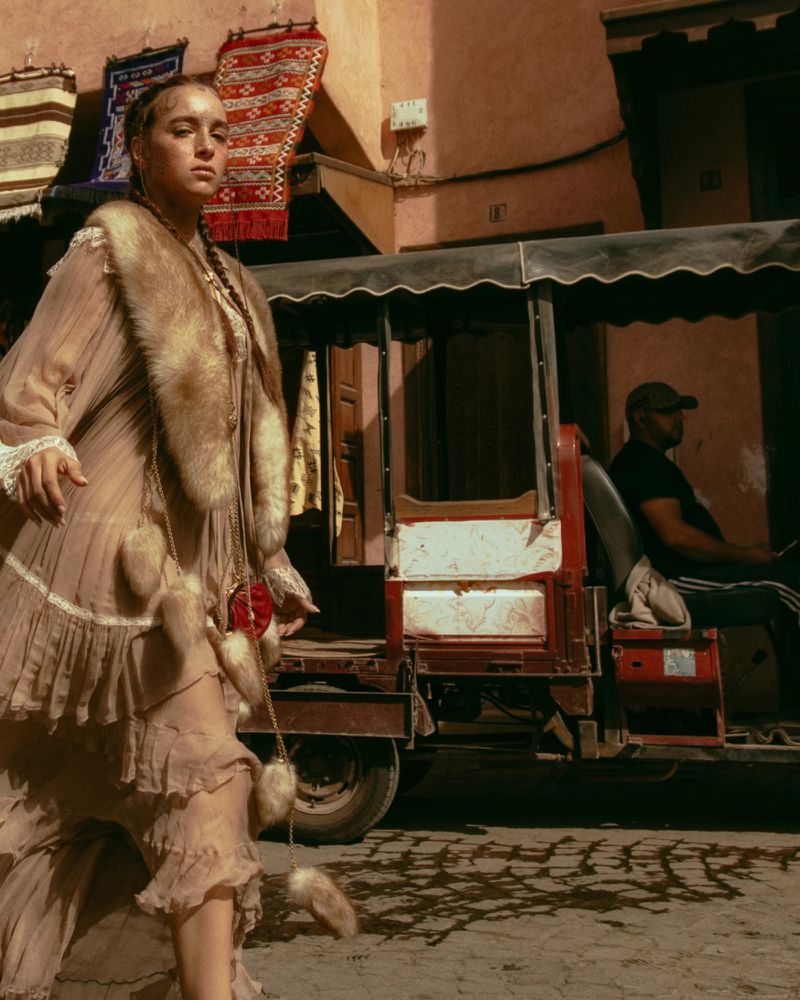 Woman in a light brown dress and fur stole walks in an outdoor urban market setting, with a passenger cart and textiles visible.