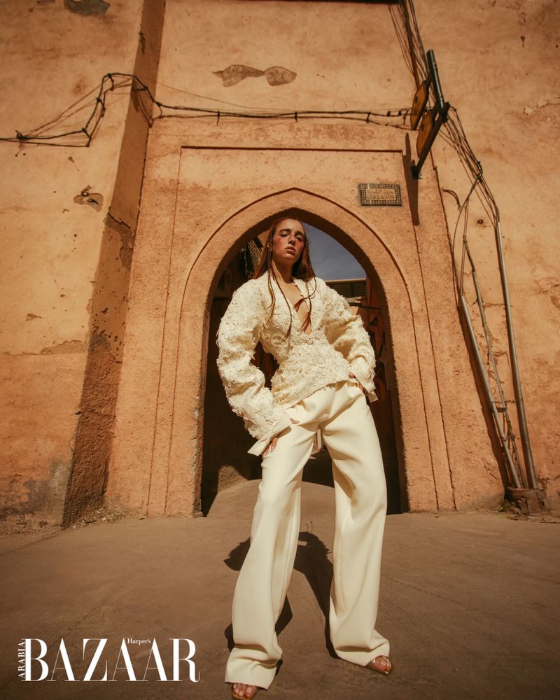 Fashion shot of a woman in a cream-colored outfit standing in an archway in an alleyway in Morocco with Harper's Bazaar logo visible.