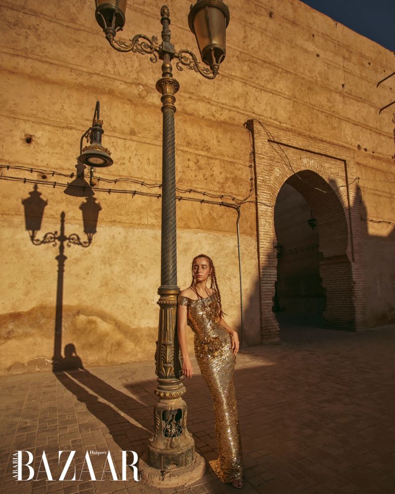 A woman in a gold dress poses in front of a vintage street lamp and an old building with an arched doorway in the background.