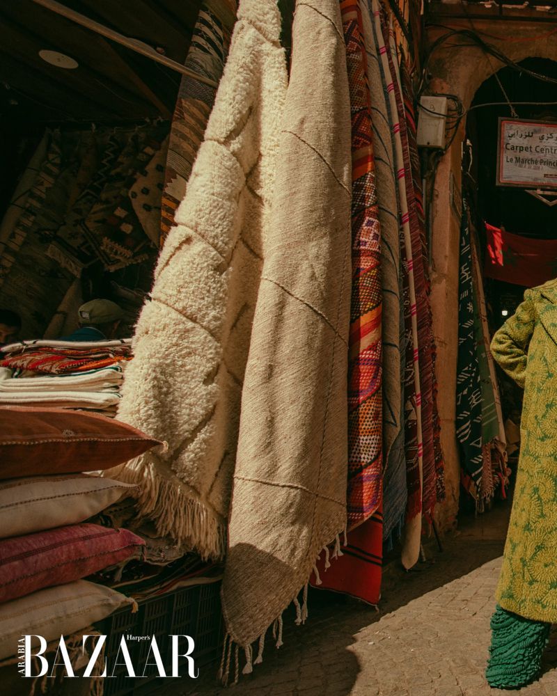 Display of artisanal rugs and pillows in a warm, earthy-toned outdoor market setting with a woman in a green patterned coat.