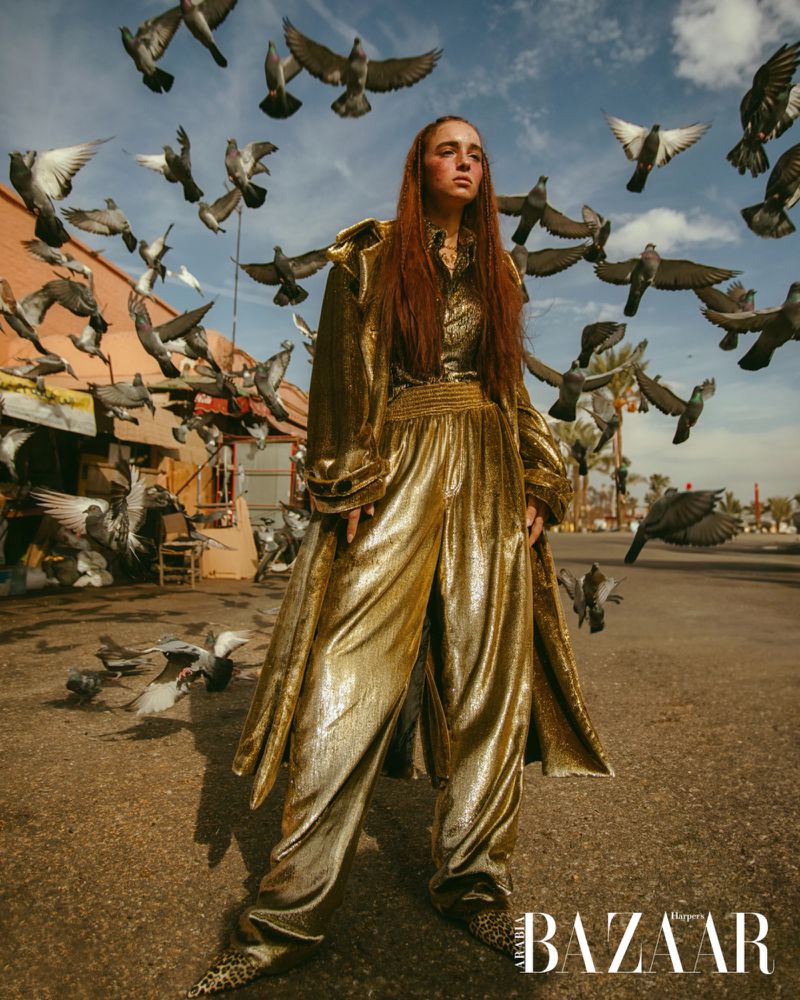 Fashion shot of a woman wearing a gold pantsuit standing in a city street surrounded by flying pigeons, posing for the camera.