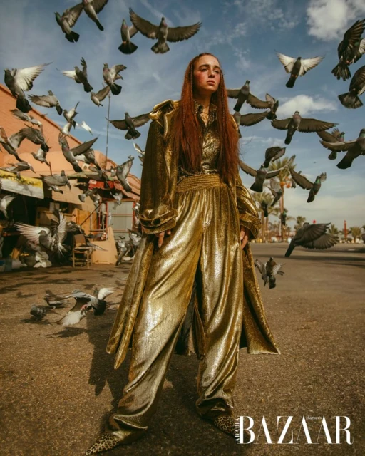 Fashion shot of a woman wearing a gold pantsuit standing in a city street surrounded by flying pigeons, posing for the camera.