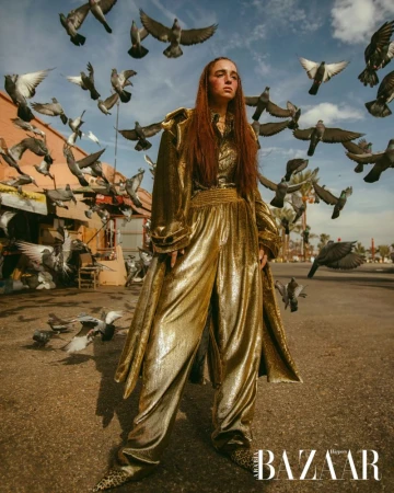 Fashion shot of a woman wearing a gold pantsuit standing in a city street surrounded by flying pigeons, posing for the camera.