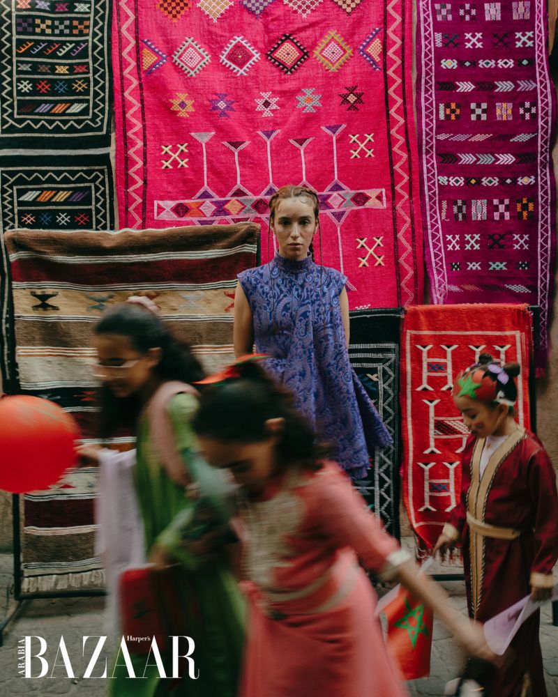 Fashion editorial shot of a model in a purple dress standing in front of colorful patterned rugs with children in the foreground.