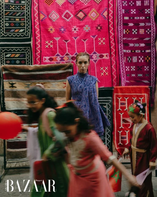Fashion editorial shot of a model in a purple dress standing in front of colorful patterned rugs with children in the foreground.