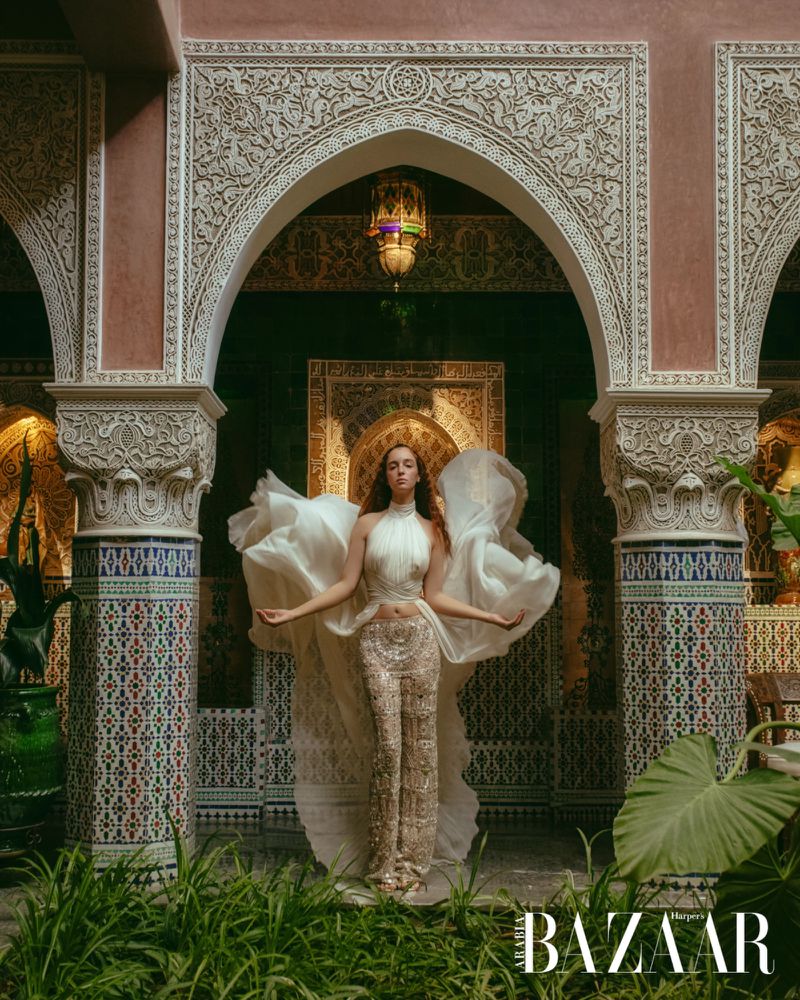A woman in a decorative outfit poses in a Moroccan-style building with arches, tilework, and plants in the foreground for a fashion editorial.