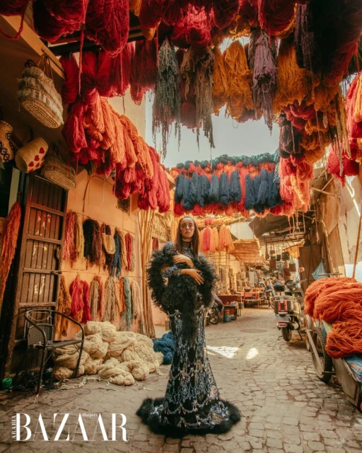 A fashion editorial shot of a woman in a dark feathered gown, standing in a narrow street in Marrakech, with colorful hanging yarn.