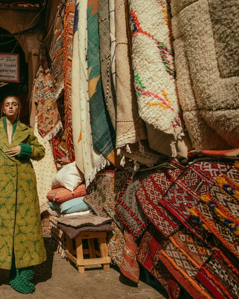 A woman in a green coat stands next to a colorful rug display at an outdoor Moroccan market, showcasing fashion and local craftsmanship.