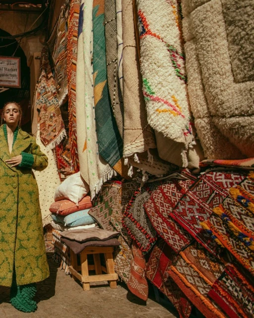 A woman in a green coat stands next to a colorful rug display at an outdoor Moroccan market, showcasing fashion and local craftsmanship.