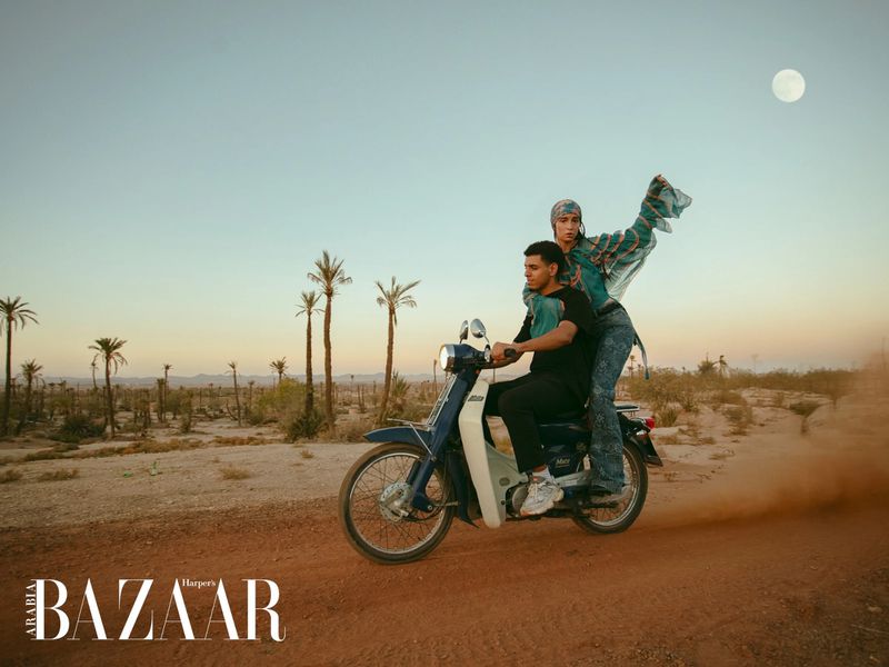 Two people ride a vintage blue motorcycle across a desert landscape beneath a bright sky, showcasing a fashion editorial for Harper's Bazaar.