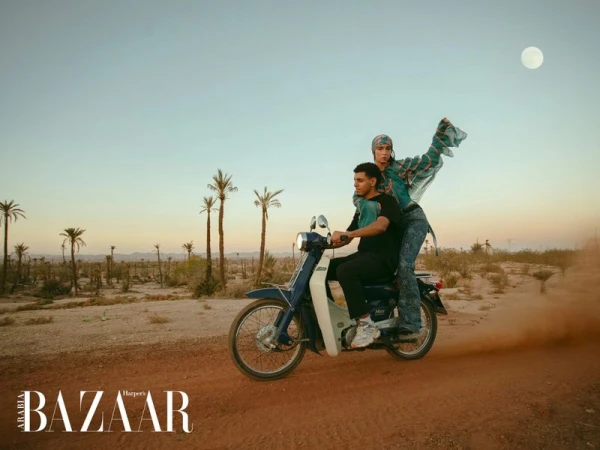 Two people ride a vintage blue motorcycle across a desert landscape beneath a bright sky, showcasing a fashion editorial for Harper's Bazaar.