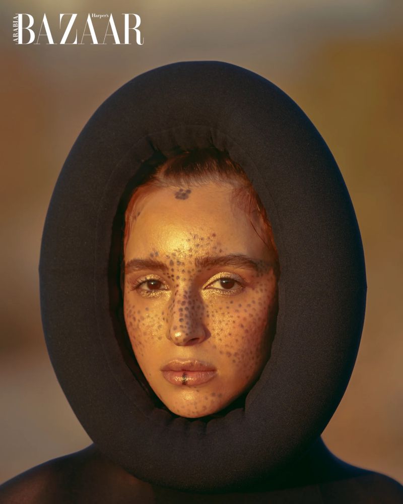 Portrait of a model with makeup and a dark hooded garment, highlighted against a neutral backdrop in a studio setting. Bazaar logo visible.