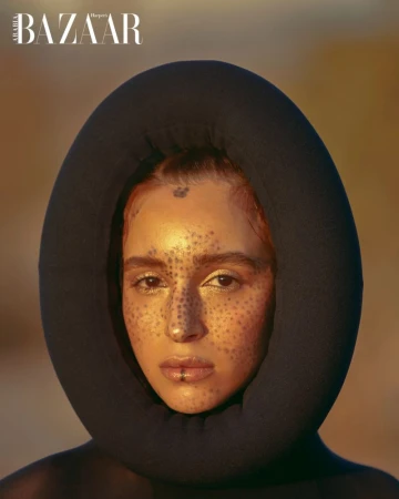 Portrait of a model with makeup and a dark hooded garment, highlighted against a neutral backdrop in a studio setting. Bazaar logo visible.