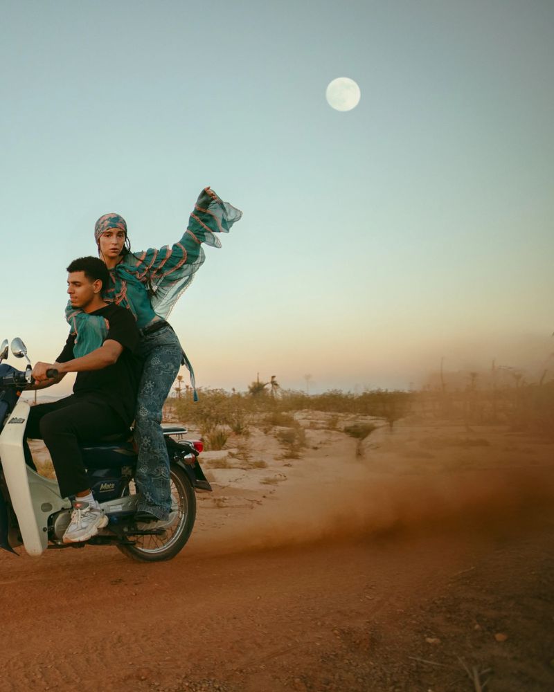 Two people ride a vintage motorcycle across a dusty landscape under a bright moon, styled in flowing garments and a headscarf.