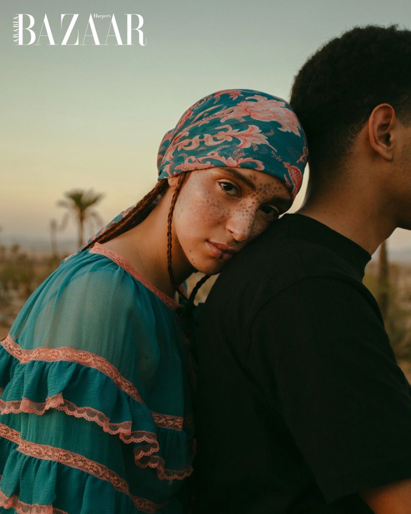 A woman rests her head on a man's shoulder, set against a backdrop of palm trees and a soft sky at dusk; fashion editorial.