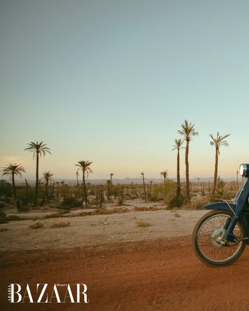A blue moped is parked on a dirt road in a desert landscape dotted with palm trees under a clear sky, as seen in a fashion editorial.