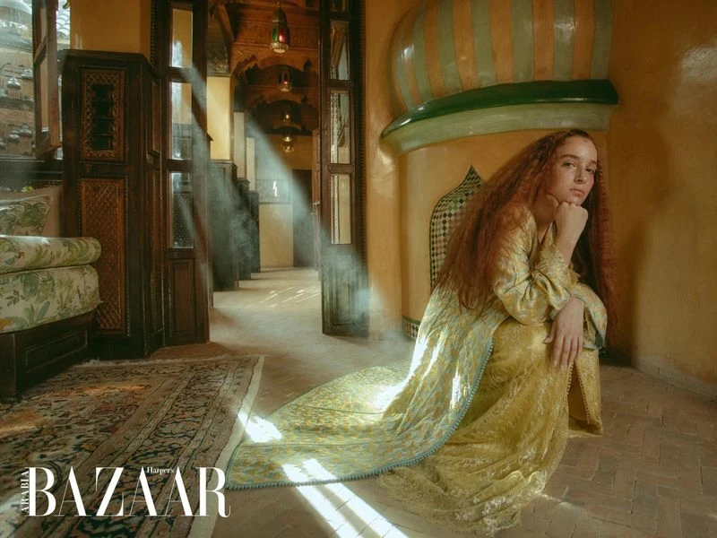 Adult woman in traditional dress poses indoors, next to doorway and archway. Light streams through the open doorway. BAZAAR logo visible.