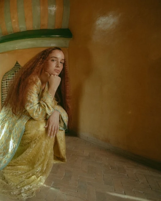 Portrait of a redhead woman in an ornate room, wearing a gold-patterned dress, seated with hand on chin, contemplative expression.