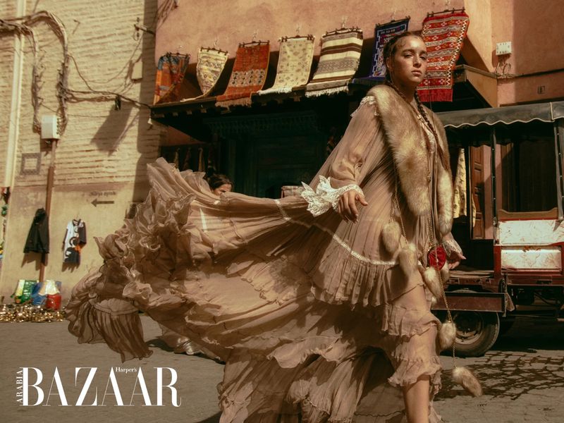 Fashion editorial shot of a woman in a long, flowing gown and fur stole, walking down a Moroccan street with traditional architecture and colors.